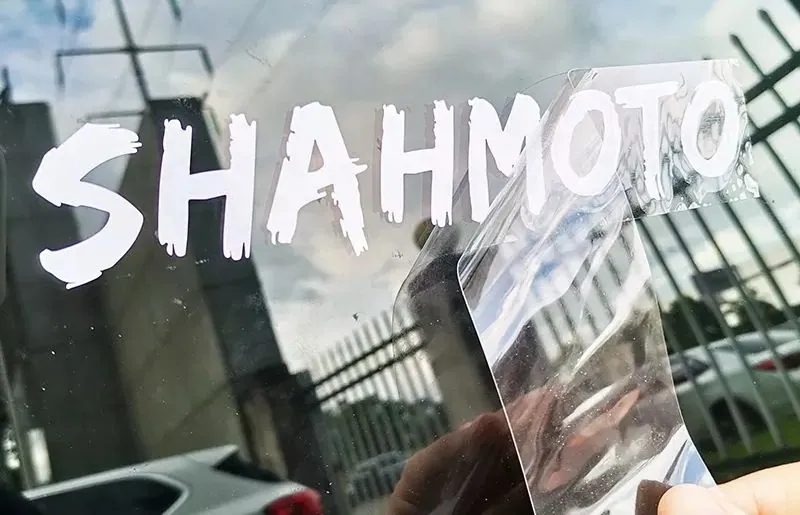 Lettrage « SHAHMOTO » en vinyle adhésif blanc posé sur une vitre de voiture, tape de transfert retiré
