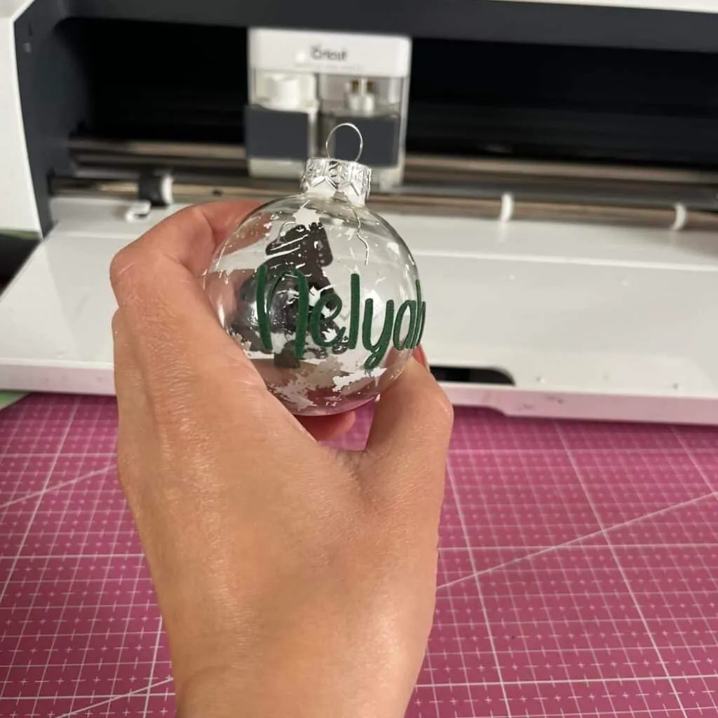 Main tenant une boule de Noël transparente sur laquelle le prénom Nelyah en vinyle vert vient d'être posé